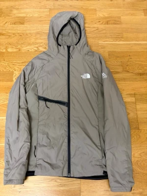 North face vindjacka - Snygg beige vindjacka från The North Face med huva och hel dragkedja framtill. Jackan har en sned bröstficka med dragkedja och diskret logga på bröstet. Tillverkad i lätt och slitstarkt material, perfekt för blåsiga dagar.