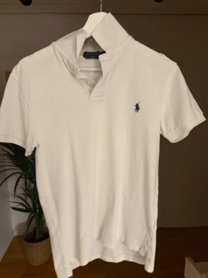 Vit piké från Polo Ralph Lauren - Klassisk vit pikétröja från Polo Ralph Lauren i slim fit-modell. Tröjan har korta ärmar, krage med två knappar och den ikoniska blå loggan broderad på bröstet. Tillverkad i mjuk bomull som känns skön mot huden.
