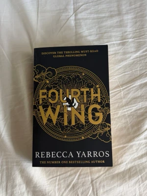 Fourth Wing av Rebecca Yarros - En populär fantasybok med svart och guld omslag, skriven av Rebecca Yarros. Boken har en stilren design med stora guldfärgade bokstäver och dekorativa detaljer. Perfekt för dig som gillar spännande och episka berättelser.