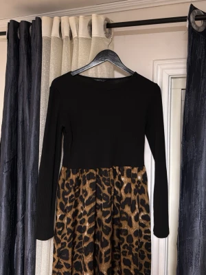 Svart långärmad klänning med leopardkjol - Snygg klänning med svart, ribbad överdel och långärmade ärmar. Kjolen har ett djärvt leopardmönster i bruna och svarta toner. Perfekt för dig som vill sticka ut med en trendig look.