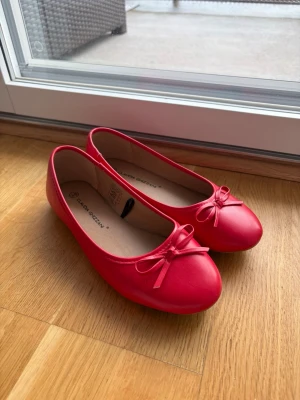 Röda ballerinaskor med rosett - Snygga röda ballerinaskor i ett glansigt material med rund tå och en söt liten rosett framtill. Skorna har en klassisk och enkel design som passar till många olika outfits. Perfekta för dig som vill ha något färgglatt och stilrent. (Endast prövade hemma och i butik)