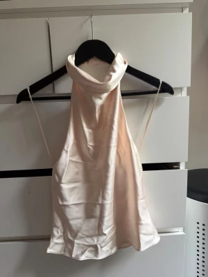 Champagnefärgad halterneck topp i satin - Snygg champagnefärgad halterneck topp i satin med hög krage och öppen rygg. Toppen har tunna axelband och ett glansigt, mjukt material som ger en lyxig känsla. 