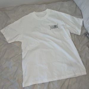 Maison margiela t-shirt - Säljer en vit oversized t-shirt med svart grafiskt tryck både fram och bak. T-shirten har korta ärmar och en klassisk rund halsringning. Perfekt för dig som gillar minimalistisk och stilren design.