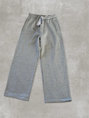 Gråa vida mjukisbyxor med dragsko - Säljer ett par gråa mjukisbyxor med breda ben och elastisk midja med dragsko. Byxorna har fickor fram och en bakficka. Tillverkade i mjukt sweatshirtmaterial som är perfekt för chill dagar. Enkla att matcha med hoodie eller t-shirt.