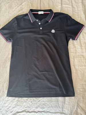 Svart pikétröja från Moncler - Klassisk svart pikétröja från Moncler med vit och röd rand på krage och ärmslut. Tröjan har knappslå med tre knappar och Moncler-logga broderad på bröstet. Perfekt till en stilren och sportig look.