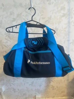 Svart och blå träningsväska Peak Performance - Snygg svart träningsväska från Peak Performance med starka blå detaljer och axelrem. Väskan har dragkedja, handtag och logga framtill. Perfekt för gymmet eller sportaktiviteter. Tillverkad i slitstarkt syntetmaterial.