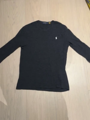 Grå långärmad tröja Polo Ralph Lauren - Stilren svart långärmad tröja från Polo Ralph Lauren med rund halsringning och den klassiska vita logotypen broderad på bröstet. Perfekt basplagg i mjukt material som passar till det mesta.