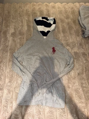 Grå hoodie med vinrött Polo Ralph Lauren-logo - Snygg grå hoodie från Polo Ralph Lauren med stor vinröd broderad logga på bröstet. Huvan har ett randigt foder i vitt och marinblått. Tillverkad i mjukt bomullsmaterial och har en klassisk passform med långa ärmar och känguruficka framtill.
