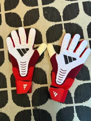 Adidas Predator målvakthandskar - Säljer ett par sportiga målvaktshandskar i rött och vitt med svarta detaljer. Handskarna har breda kardborreband vid handleden och greppvänlig insida. Perfekta för dig som gillar fotboll och vill ha bra grepp i målet.
