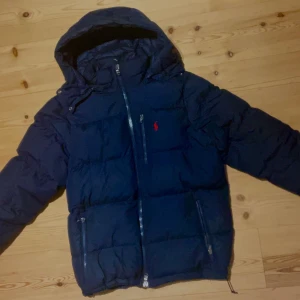 Mörkblå Ralph Lauren pufferjacka med huva - Snygg mörkblå pufferjacka med huva och dragkedja framtill. Jackan har två sidofickor med dragkedja och en bröstficka med dragkedja den har även en till ficka innanför jackan. En röd Ralph Lauren logga på bröstet. Perfekt för kalla vinterdagar.
