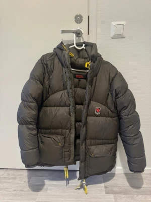 Mörkgrön pufferjacka från Fjällräven - Säljer en mörkgrön pufferjacka från Fjällräven med huva och gula detaljer på snörningarna. Jackan har två stora fickor framtill med knappar och klassisk Fjällräven-logga på bröstet. Perfekt för kalla vinterdagar och riktigt skön att bära.