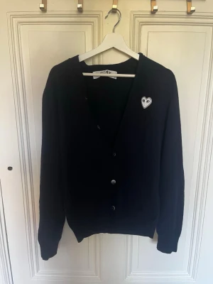 Mörkblå cardigan Comme des Garçons Play - Mörkblå cardigan från Comme des Garçons Play med klassisk passform och knäppning framtill. Ikoniskt hjärta med ögon broderat på bröstet. Mjuk och skön, perfekt att slänga på sig över en t-shirt. Stilren och enkel design.