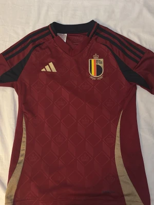 matchtröja i vinrött från Adidas - Snygg vinröd Belgien matchtröja från Adidas med guldiga detaljer och svarta ränder på axlarna. Tröjan har Belgiska fotbollsförbundets emblem på bröstet och är tillverkad i ett lätt, ventilerande material. Perfekt för fotbollsträning eller att bära till match.