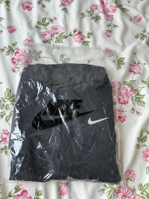 Svart Nike vindjacka med huva - Snygg svart vindjacka från Nike med dragkedja framtill och huva. Jackan har Nike-logga i vitt på bröstet och är tillverkad i lätt, vindtätt material. Perfekt för blåsiga dagar och sportiga outfits.