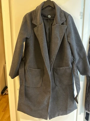 Mörkgrå ullrock med stora fickor - Snygg mörkgrå ullrock från Jole Moda med breda slag, två stora utanpåfickor och knytskärp i midjan. Rocken har en rak passform och helsvart foder. Perfekt för kyliga dagar när du vill vara både varm och stilren.