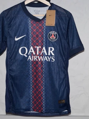 PSG matchtröja blå Nike - Snygg blå Paris Saint-Germain matchtröja från Nike med röd och vit mönstrad rand i mitten. Tröjan har klubbmärke på bröstet, vit Nike-logga och Qatar Airways som sponsortryck. Perfekt för fotbollsträning eller att bära på läktaren. Är du efter en annan storlek är det bara att skriva!