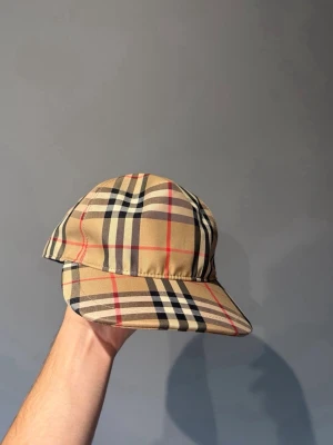 Burberry keps  - Säljer en klassisk beige keps från Burberry med det ikoniska rutmönstret i svart, vitt och rött. Kepsen har justerbar rem bak och en patch med texten 'Burberry London England'. Perfekt accessoar för en trendig look.