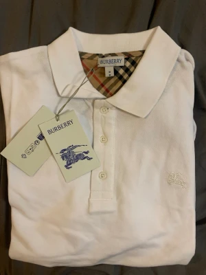 Vit pikétröja från Burberry - Klassisk vit pikétröja från Burberry med den ikoniska broderade loggan på bröstet och märkets rutiga detalj i kragen. Tröjan har krage, knappar framtill och är tillverkad i ett strukturerat bomullstyg. Perfekt för en stilren och tidlös look.