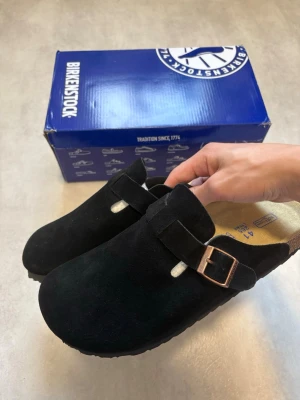 Birkenstock Boston Svarta Mocka Strl 41 - Säljer ett par svarta Birkenstock Boston i storlek 41. Endast provade en gång inomhus och är i mycket fint skick utan några brister.  Klassisk och bekväm modell i mocka med justerbart metallspänne och fotbädd i kork. Perfekta både hemma och ute!  Vid bud eller funderingar är det bara att skriva! 😊