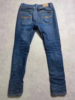 Blå slim fit jeans från Nudie Jeans - Snygga blå slim fit jeans från Nudie Jeans med klassiska fem fickor och kontrastsömmar. Jeansen har en rak passform, läderpatch bak i midjan och signaturbroderi på bakfickorna. Tillverkade i slitstark denim av bomull.