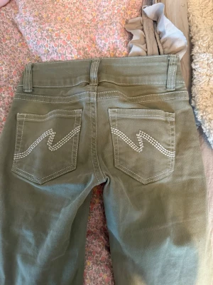 Olivgröna jeans från Nelly - Snygga olivgröna jeans från Nelly med raka ben och klassisk femficksmodell. Bakfickorna har dekorativa vita sömmar i zigzag-mönster. Jeansen har bälteshällor och stängs med knapp och dragkedja. Perfekta för en trendig och avslappnad stil.
