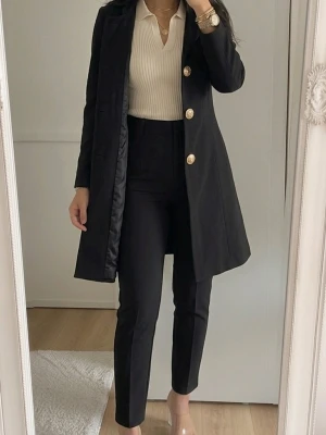 Svart rock med guldknappar - Stilren svart rock med raka linjer och fyra stora guldknappar framtill. Rocken har en klassisk krage och är knälång med figurnära passform. Perfekt för dig som gillar en elegant och tidlös look. 
