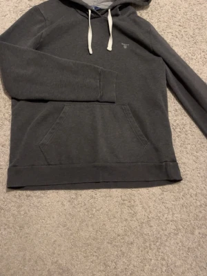 Mörkgrå hoodie från GANT - Snygg mörkgrå hoodie från GANT med vit dragsko i huvan och klassisk känguruficka framtill. Diskret GANT-logga broderad på bröstet. Tillverkad i mjukt bomullsmaterial och har långa ärmar. Perfekt för en chill och avslappnad stil.