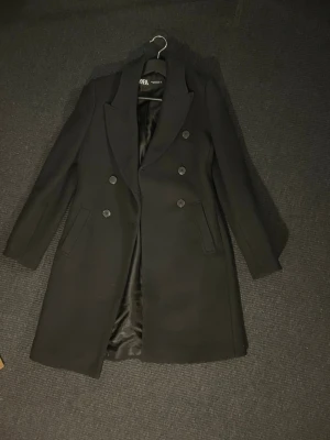 Svart dubbelknäppt kappa - Stilren svart kappa med dubbelknäppning och klassisk krage. Kappan har två fickor framtill och är helfodrad med slät insida. Perfekt för en trendig och tidlös look under kallare dagar.