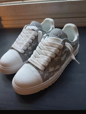 Gråvita sneakers från Numeris - Snygga sneakers från Numeris i grått och vitt med camomönster. Skorna har tjock vit sula, vita skosnören och tåparti i texturerat material. Perfekta för dig som gillar streetstyle och vill sticka ut lite extra.