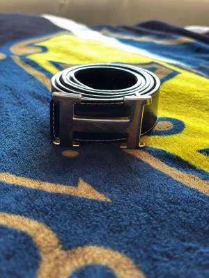 Svart skärp med metallspänne - Snyggt svart skärp i skinn med ett stort H-format metallspänne. Skärpet har vita sömmar längs kanterna som ger en schysst kontrast. Perfekt accessoar för att lyfta din outfit.