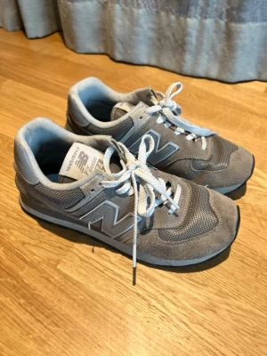 Grå New Balance sneakers - Klassiska grå sneakers från New Balance med stora N-loggor på sidorna. Skorna har ovandel i mocka och mesh, vita skosnören och en slitstark gummisula med bra grepp. Perfekta för dig som gillar en sportig och stilren look.