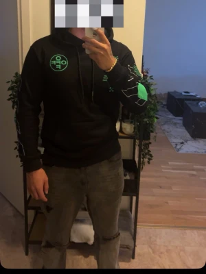 Svart hoodie med grönt tryck - Svart hoodie med huva och gröna grafiska tryck samt textdetaljer på både framsidan och ärmen. Stora Off-White-inspirerade motiv på ryggen och bröstet. Känguruficka framtill och snören i huvan.