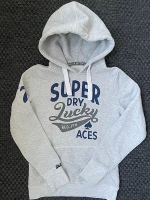 Grå hoodie med tryck från Superdry - Säljer nu denna sjukt snygga Vintage 2000s SuperDry hoodie! Passar till allt och har en perfekt passform. Säljs ej längre i butik och är därav mycket eftertraktad😊 passar xs perfekt ! Skriv vid funderingar 🤝