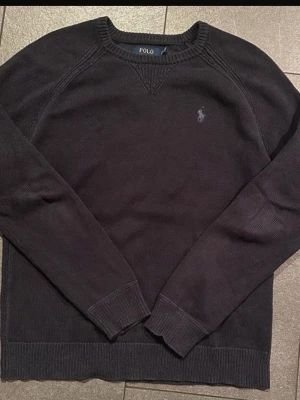 Mörkblå stickad tröja Polo Ralph Lauren - Mörkblå stickad tröja från Polo Ralph Lauren med rund halsringning och diskret broderad logga på bröstet. Tröjan har långa ärmar och ribbade muddar vid ärmslut och nederkant. Perfekt till en clean och klassisk stil.