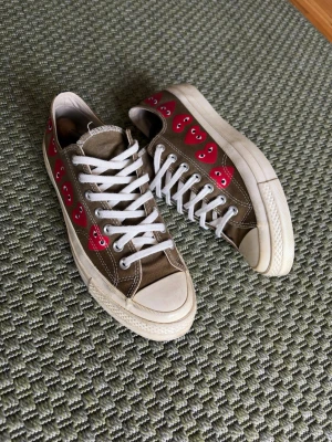 Bruna sneakers med röda hjärtan - Säljer ett par bruna låga sneakers med vita sulor och snören. Skorna har flera röda hjärtan med ögon längs sidorna som ger en unik och lekfull look. Tillverkade i canvasmaterial och har klassisk tåhätta i gummi. Perfekta för dig som vill sticka ut.