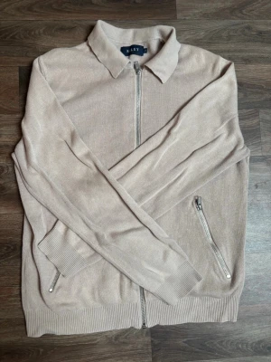 Beige ziptröja från Riley - Stilren beige ziptröja från Riley med krage och hel dragkedja framtill. Tröjan har två fickor med dragkedja och ribbade muddar vid ärmslut och nederkant. Perfekt för en avslappnad men snygg look.