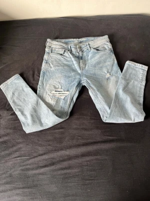 Ljusa slitna jeans - Snygga ljusblå jeans med slitningar och hål på låret och knät. Jeansen har klassisk femficksdesign och dragkedjegylf. Perfekta för en avslappnad och trendig look.