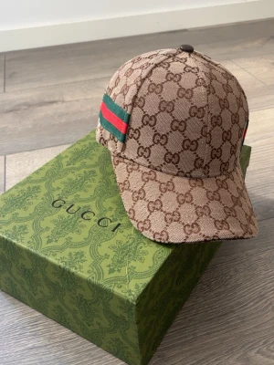 Gucci beige monogram keps - Säljer en beige keps från Gucci med klassiskt GG-monogram över hela kepsen och grönt/rött band på sidan. Kepsen har böjd skärm och justerbar rem bak. Materialet är canvas med detaljer i läder. Perfekt för dig som vill ha en ikonisk och trendig accessoar.
