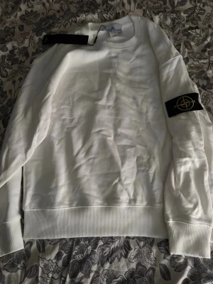 Vit sweatshirt från Stone Island - Vit sweatshirt från Stone Island med klassisk rund halsringning och ribbade muddar. Tröjan har den ikoniska svarta logotypen på vänster ärm. Tillverkad i mjukt material som känns skönt mot huden. Perfekt för en clean och stilren look.