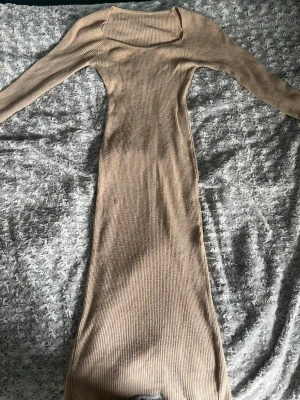 Beige ribbad långklänning - Säljer en beige ribbad långklänning med lång ärm och fyrkantig ringning. Klänningen har en figurnära passform och en slits baktill för extra rörelsefrihet. Perfekt för dig som gillar stilrena och enkla plagg med en modern touch.