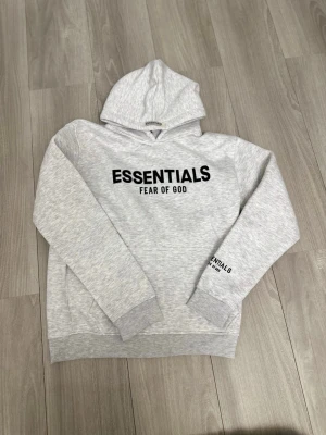 Grå Essentials Fear of God hoodie REP - Ljusgrå hoodie från Essentials Fear of God med svart tryck på bröstet och ärmen. Klassisk passform med huva och ribbade muddar vid ärmslut och nederkant. Tillverkad i mjukt sweatshirtmaterial som känns skönt mot huden.