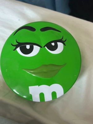 Grön M&M's plåtburk - Säljer en rund grön plåtburk med motiv av den gröna M&M-karaktären. Burken har lock och är dekorerad med ansikte och ett stort vitt 'm' på framsidan. Perfekt för förvaring av småsaker eller godis. Har lite repor. Från 2015. 