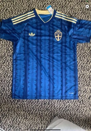 Blå Sverige fotbollströja Adidas - Säljer en blå fotbollströja från Adidas med Sveriges landslagsemblem på bröstet. Tröjan har gula detaljer, klassiska Adidas-ränder på axlarna och ett mönster i olika blå nyanser. Tillverkad i lätt och ventilerande material, perfekt för träning eller match.