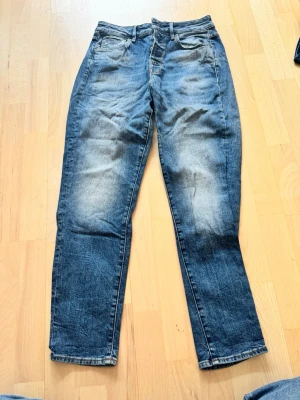 G-star raka jeans 27/30 - Säljer ett par blå jeans med raka ben och snyggt slitna detaljer. Jeansen har klassisk femficksmodell och knappgylf framtill. Materialet är denim i bomull och färgen är mellanblå med ljusare partier på låren och knäna.