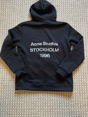 Svart hoodie Acne Studios Stockholm - Snygg svart hoodie från Acne Studios med vit text 'Acne Studios STOCKHOLM 1996' på ryggen. Klassisk modell med huva och magficka, ribbade muddar vid ärmslut och nederkant. Perfekt för en clean och stilren look. Ni vet varför priset är som de är🔥