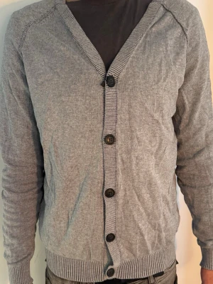 Grå cardigan från Selected Homme - Snygg grå cardigan från Selected Homme med v-ringning och fem knappar framtill. Tillverkad i mjukt stickat material och har ribbade muddar vid ärmslut och nederkant. Perfekt att slänga över en t-shirt för en avslappnad look.