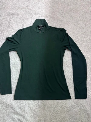 Mörkgrön polotopp från Monki - Snygg mörkgrön polotopp från Monki i mjukt material. Toppen har lång ärm och hög krage med en liten snörningsdetalj framtill. Perfekt att styla med jeans eller kjol för en clean look.