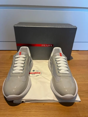 P r a d a cups skor  - Snygga gråa sneakers från P r a d a  med vita detaljer och vita skosnören. Skorna har en modern siluett med meshpaneler och glansiga partier i syntetmaterial. Röd P r a d a -logga på plösen ger en exklusiv touch. Perfekta för dig som vill ha stilrena och trendiga sneakers.