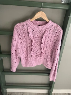 Ljuslila stickad tröja med volang - Supersöt ljuslila stickad tröja med volangdetaljer framtill och hålmönster över hela plagget. Tröjan har rund halsringning, ribbade muddar och är gjord i ett mjukt garn som känns skönt mot huden. Perfekt för dig som gillar romantiska detaljer och pastellfärger.