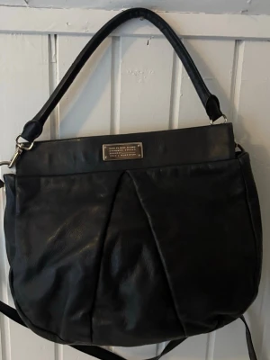 Marc Jacobs Too hot to handle väska - Snygg svart Marc Jacobs  handväska med rundad form och dekorativ metallbricka framtill. Väskan har både axelrem och handtag, vilket gör den flexibel att bära. Materialet är i äkta skinn och väskan har en stilren, enkel design som passar till det mesta.